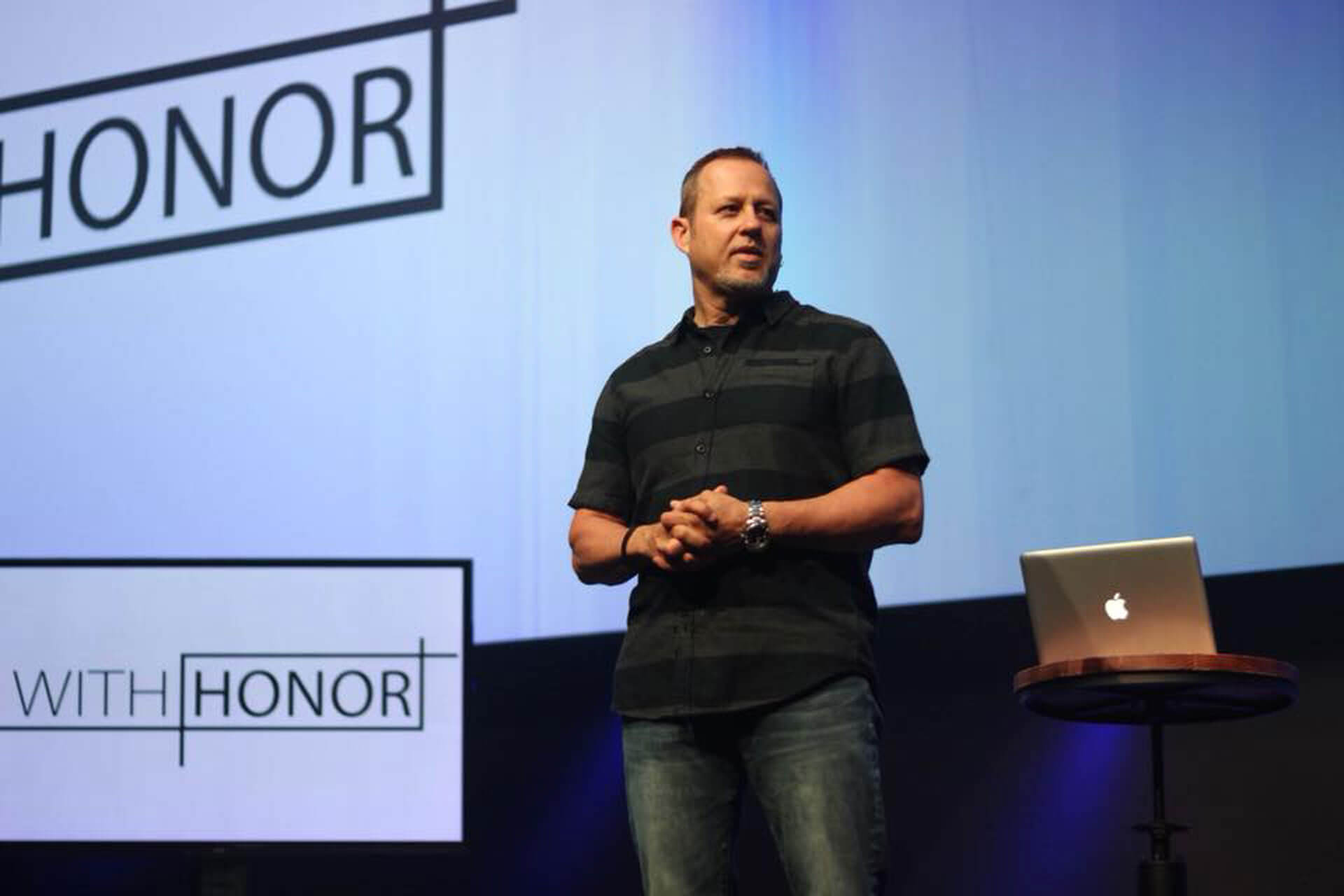 Man presenting on stage with text "with honor" on screen behind him.