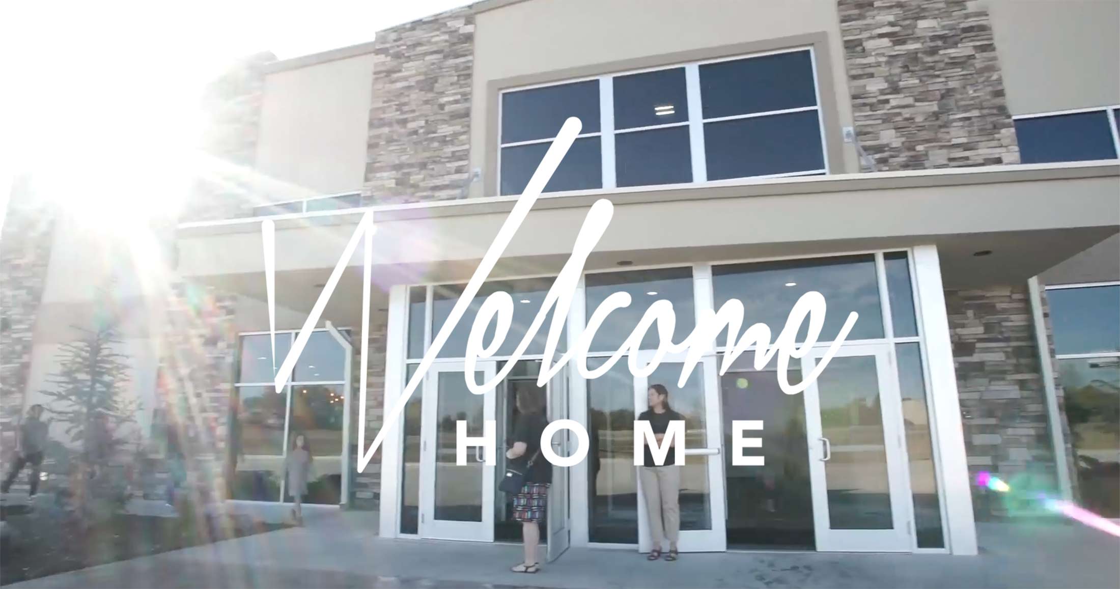 Sunlight flares over a modern building with a large "welcome home" sign on the glass door where a woman is standing outside the church in Shawnee Oklahoma