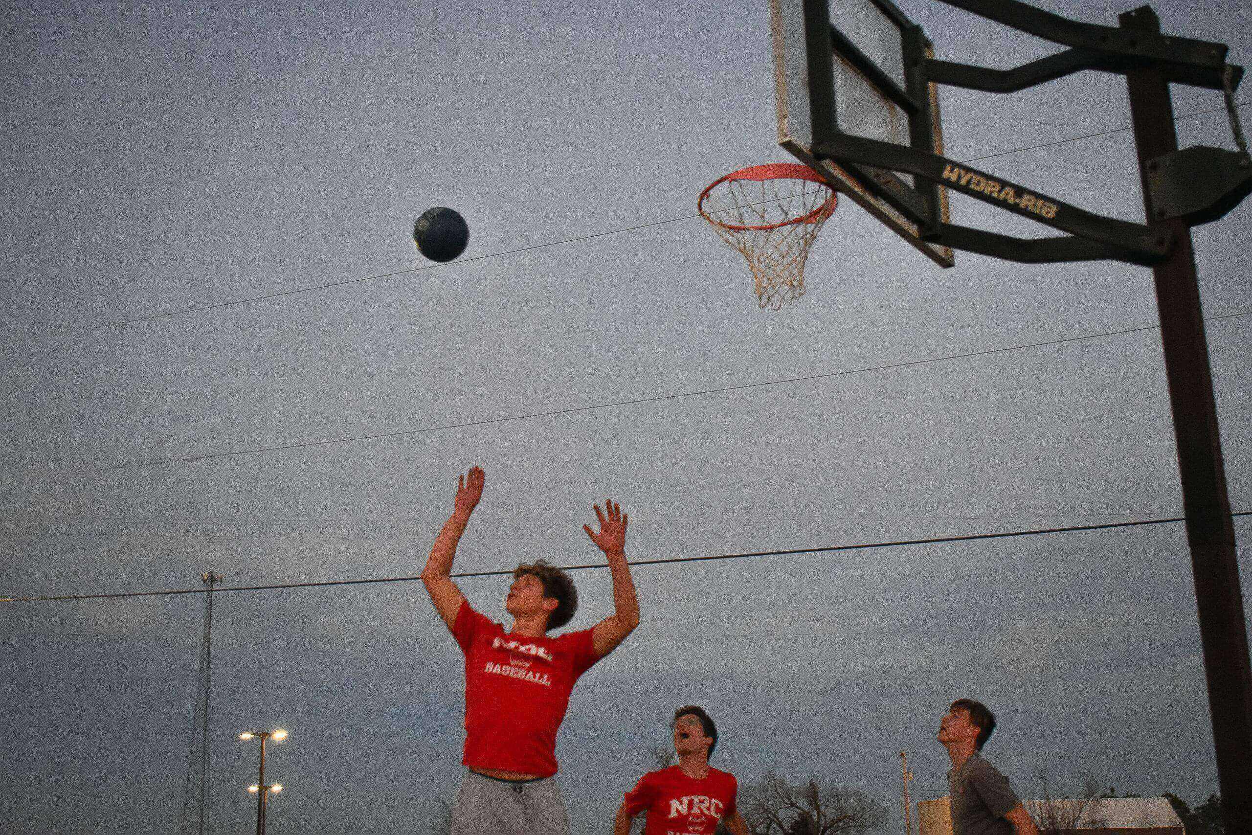 Three individuals playing basketball outdoors, one of whom is jumping to shoot the ball towards the hoop.